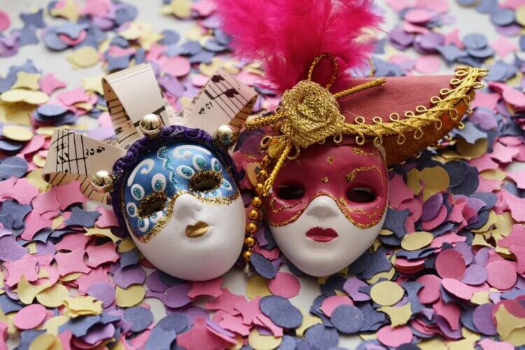 Twee Venetiaanse carnavalsmaskers op gekleurde confetti; één blauw met muzieknoten, één roze met goud en veer. Wees jezelf advies.
