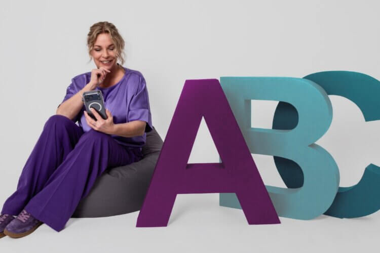 Claudia Bouwens blickt lächelnd auf ihr Telefon, neben großen bunten ABC-Buchstaben in Plum und Teal.